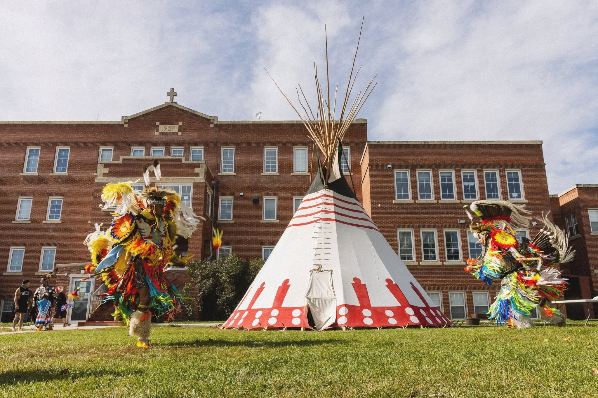 Two dancers in colourful regalia performing on the lawn beside the tipi