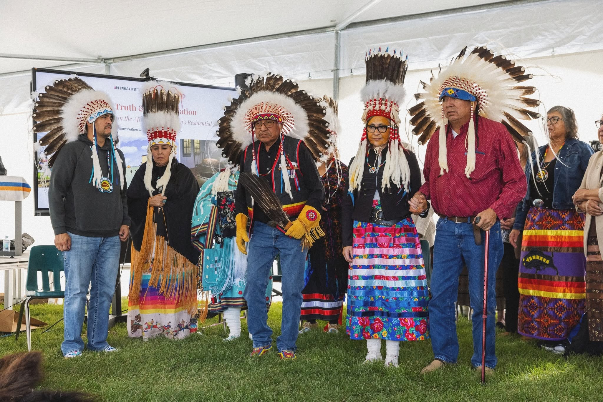 Honourees wearing feathered headdresses and ribbon skirts standing together in a tent