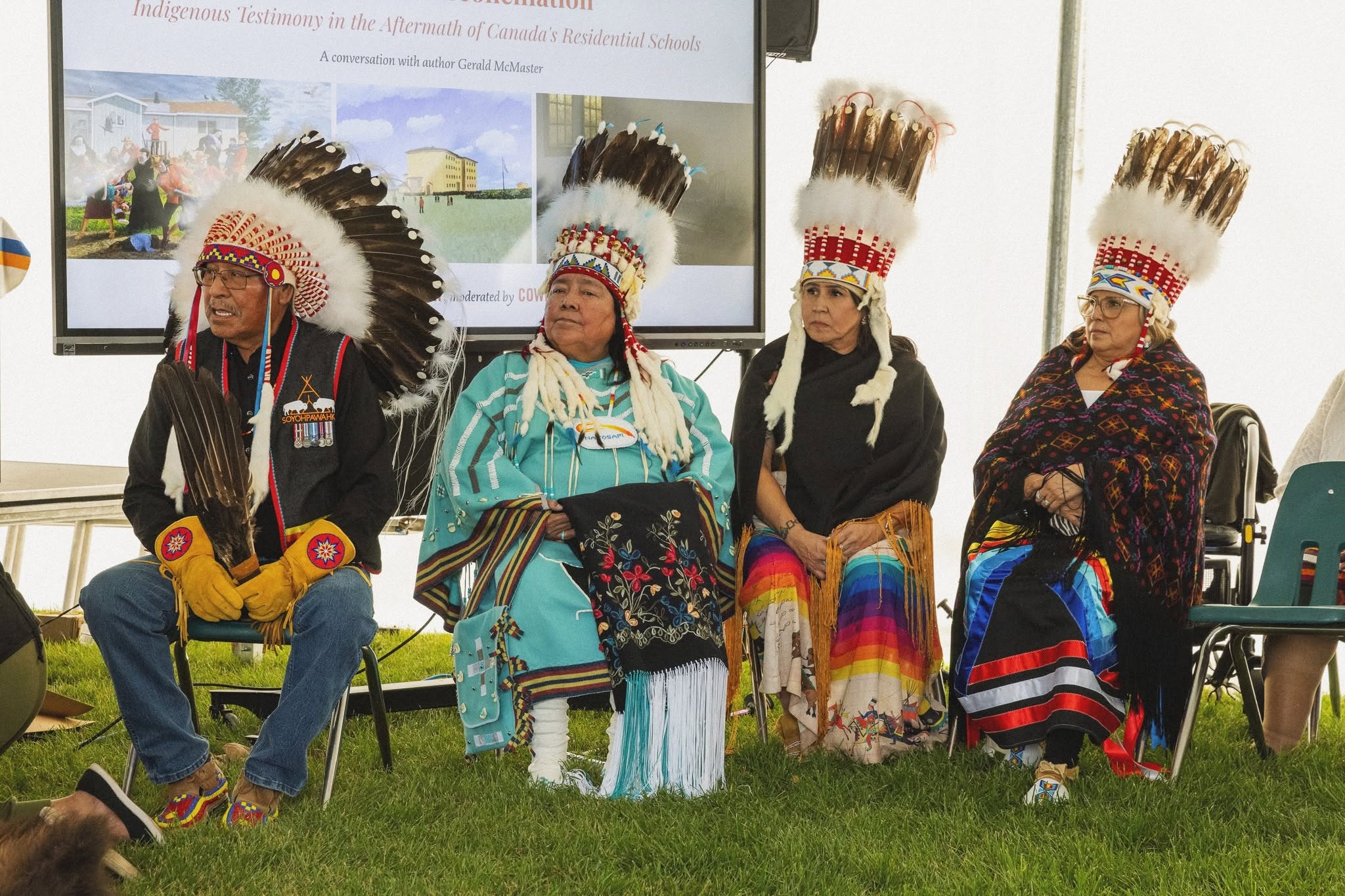 Four seated honourees in headdresses and traditional regalia in front of a presentation screen