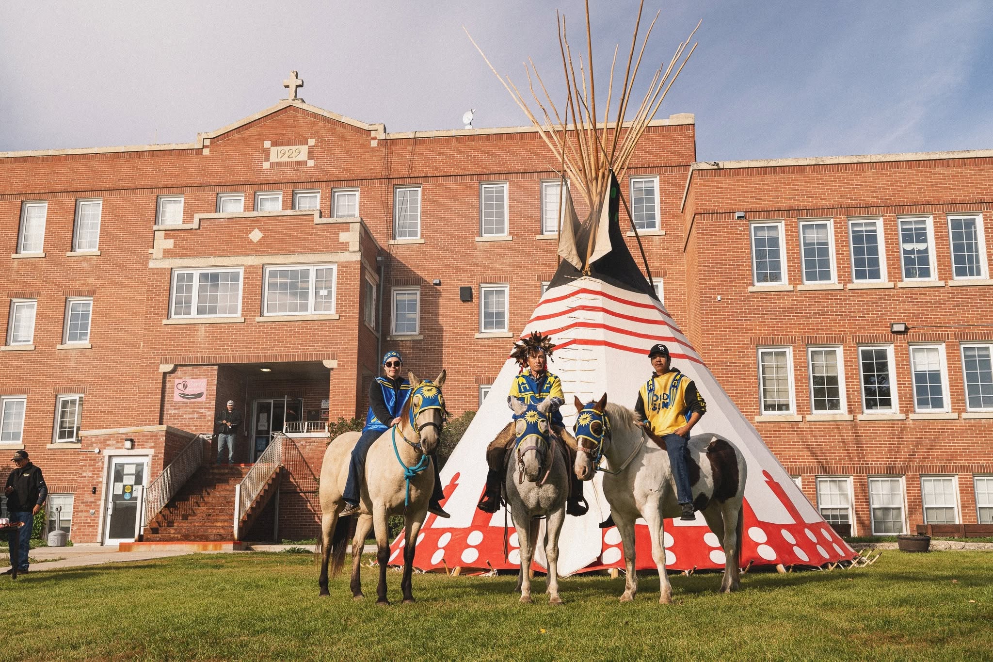 Three riders on horses wearing Old Sun colours in front of the tipi and college building